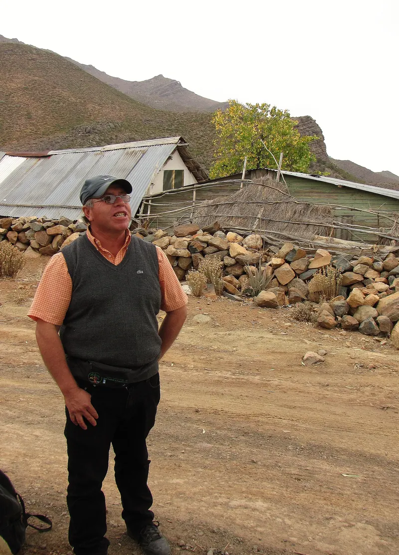 Hombre de pie frente a un muro de piedra seca y una ramada de varas, con cerros áridos detrás