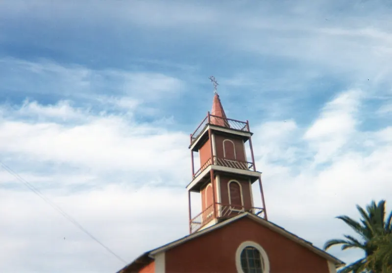 Torre de madera pintada, con balcones, chapitel y cruz, sobre un frontis rojo con un óculo
