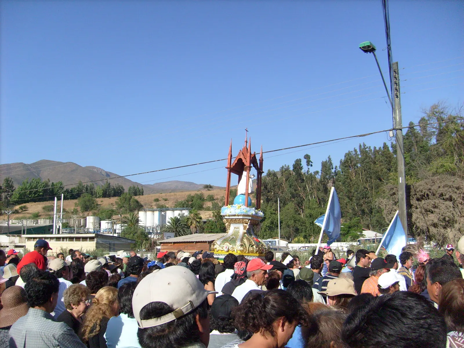Multitud con banderas celestes alrededor de un anda alzada, con cerros áridos y eucaliptos al fondo