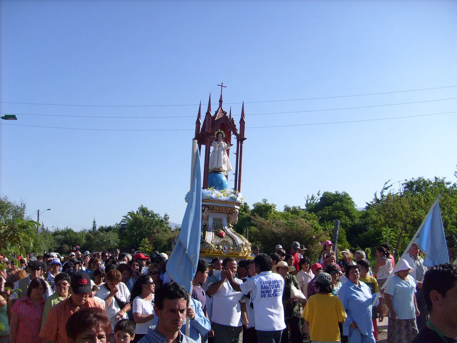 Multitud de peregrinos con banderas celestes alrededor de un anda alta de estilo neogótico con la imagen del Niño